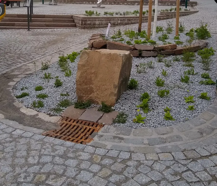 Platzfläche aus Natursteinpflaster im Bogenverband mit geschwungener Einfassung, Kiesbeet, Sandsteinblock und Entwässerungsrinne